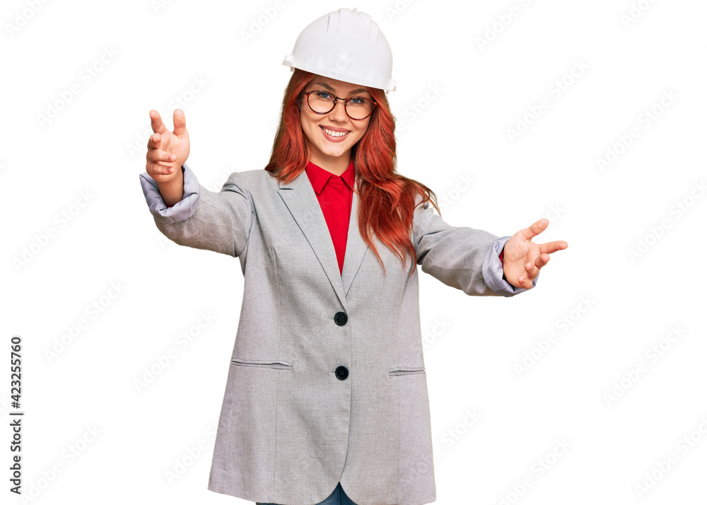 Young redhead woman wearing architect hardhat looking at the camera smiling with open arms for hug. cheerful expression embracing happiness. Young redhead woman wearing architect hardhat looking at the camera smiling with open arms for hug. cheerful expression embracing happiness.