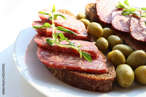 Close-up of Salami pepperoni on rye bread sandwich with green pickled olives on plate on white table background. Flat lay food. Top view, copy space. Selective focus