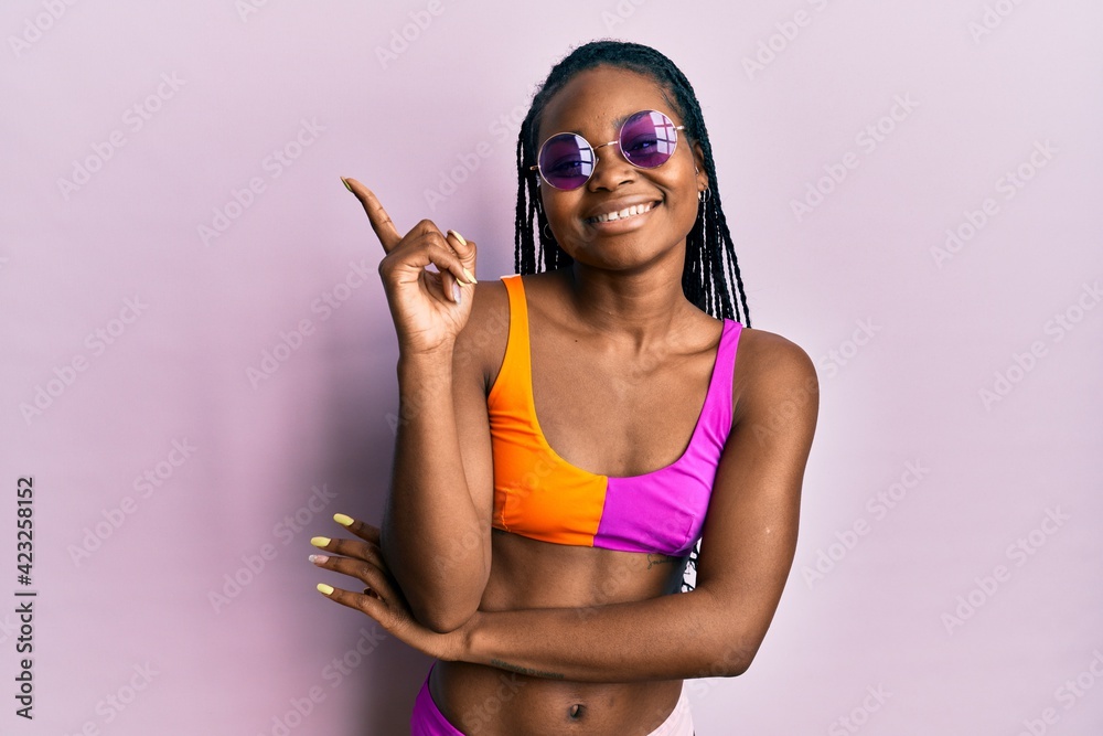 Young african american woman wearing bikini and sunglasses with a big smile on face, pointing with hand and finger to the side looking at the camera.