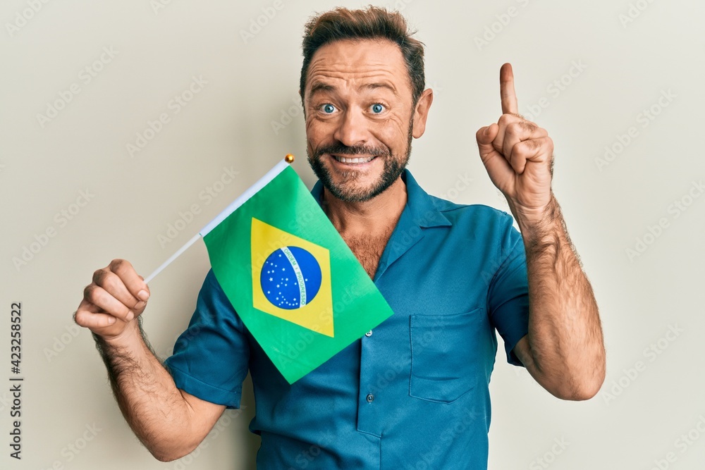 Middle age man holding brazil flag smiling with an idea or question pointing finger with happy face, number one Middle age man holding brazil flag smiling with an idea or question pointing finger with happy face, number one