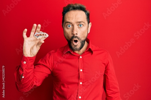 Middle age man holding brilliant diamond stone scared and amazed with open mouth for surprise, disbelief face