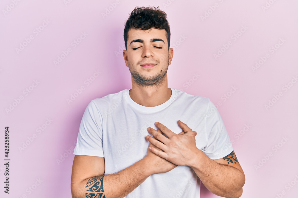 Young hispanic man wearing casual white t shirt smiling with hands on chest, eyes closed with grateful gesture on face. health concept. Young hispanic man wearing casual white t shirt smiling with hands on chest, eyes closed with grateful gesture on face. health concept.