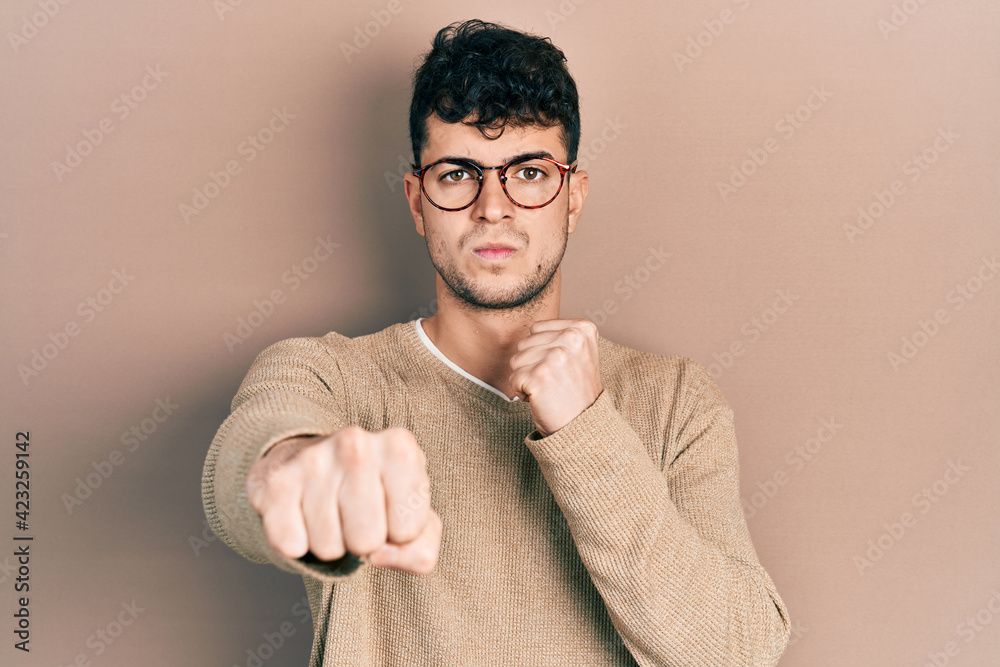 Young hispanic man wearing casual clothes and glasses punching fist to fight, aggressive and angry attack, threat and violence Young hispanic man wearing casual clothes and glasses punching fist to fight, aggressive and angry attack, threat and violence