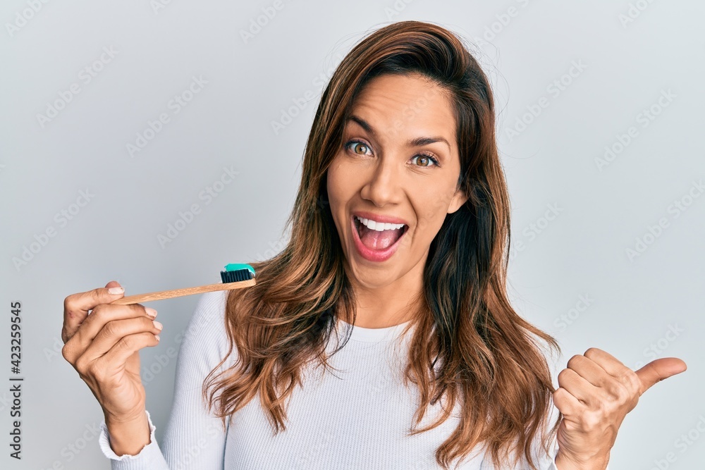 Young latin woman holding toothbrush with toothpaste pointing thumb up to the side smiling happy with open mouth Young latin woman holding toothbrush with toothpaste pointing thumb up to the side smiling happy with open mouth