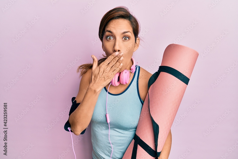 Young latin woman holding yoga mat covering mouth with hand, shocked and afraid for mistake. surprised expression Young latin woman holding yoga mat covering mouth with hand, shocked and afraid for mistake. surprised expression
