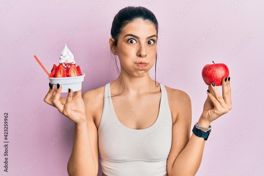 Young hispanic woman wearing sportswear holding apple and ice cream puffing cheeks with funny face. mouth inflated with air, catching air.