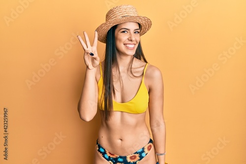 Young hispanic woman wearing bikini and summer hat showing and pointing up with fingers number three while smiling confident and happy.