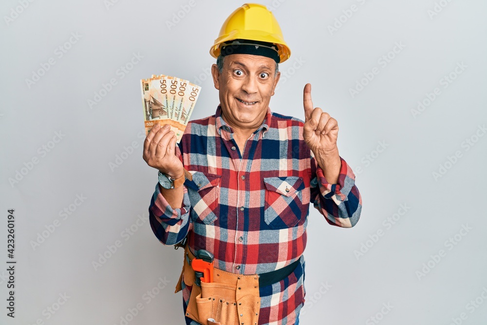 Senior hispanic man wearing handyman uniform holding norwegian krona smiling with an idea or question pointing finger with happy face, number one