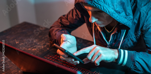 Hacker with phone is typing on a laptop keyboard in a dark room under a neon light. Cybercrime fraud and identity theft