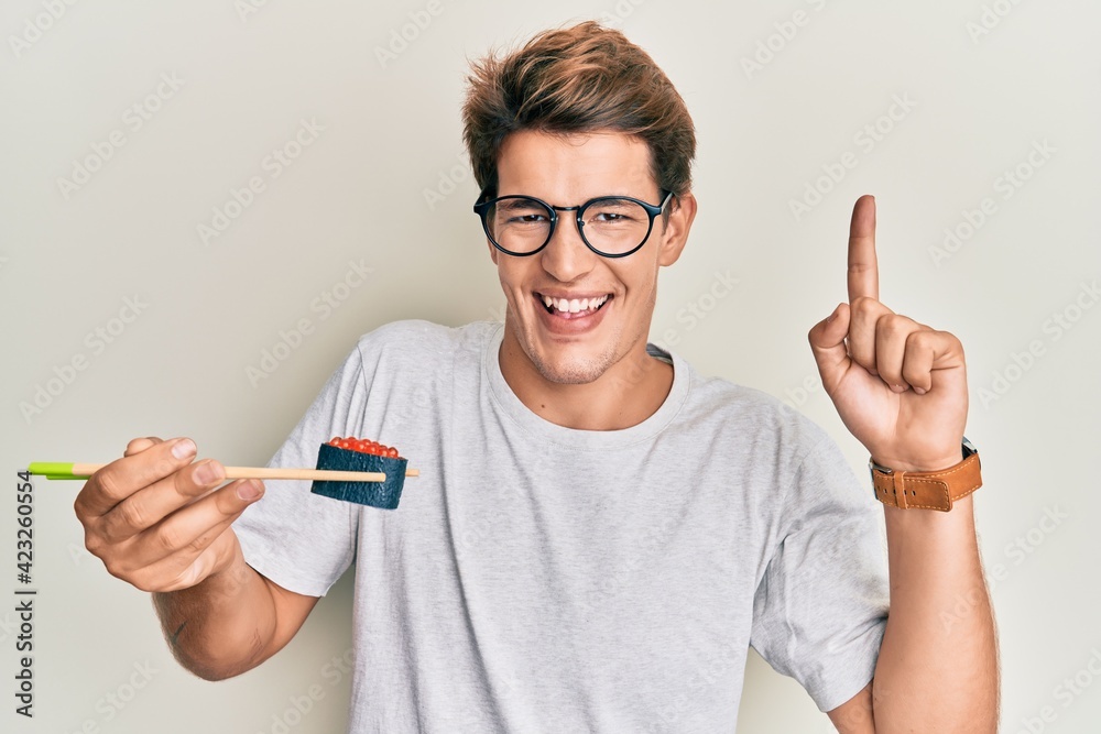 Handsome caucasian man eating sushi using chopsticks smiling with an idea or question pointing finger with happy face, number one