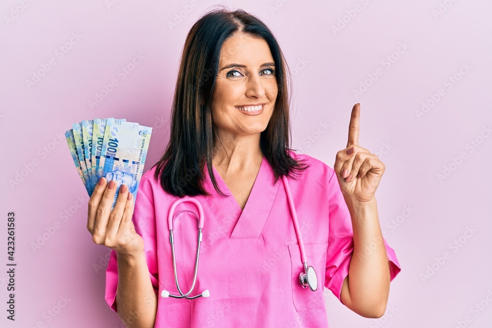 Middle age brunette doctor woman holding 100 south african rand banknotes smiling with an idea or question pointing finger with happy face, number one Middle age brunette doctor woman holding 100 south african rand banknotes smiling with an idea or question pointing finger with happy face, number one