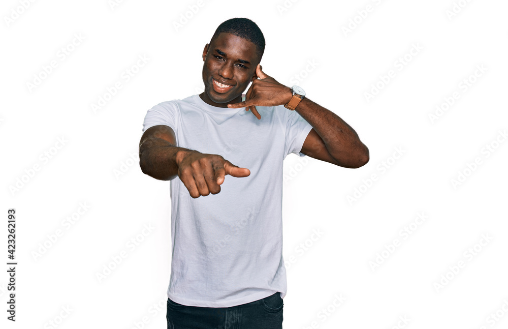 Young african american man wearing casual white t shirt smiling doing talking on the telephone gesture and pointing to you. call me.