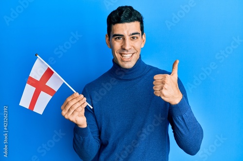Handsome hispanic man holding england flag smiling happy and positive, thumb up doing excellent and approval sign