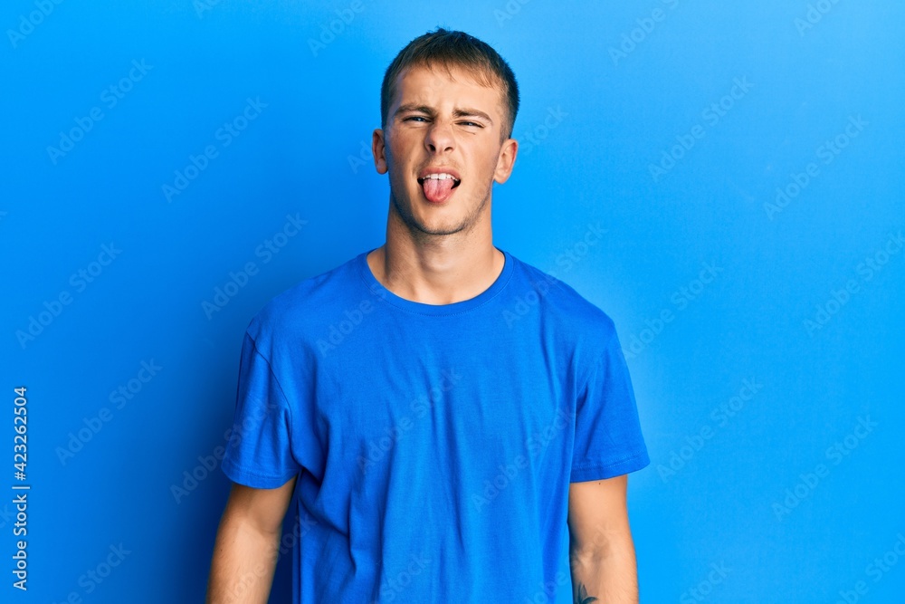 Young caucasian man wearing casual blue t shirt sticking tongue out happy with funny expression. emotion concept. Young caucasian man wearing casual blue t shirt sticking tongue out happy with funny expression. emotion concept.