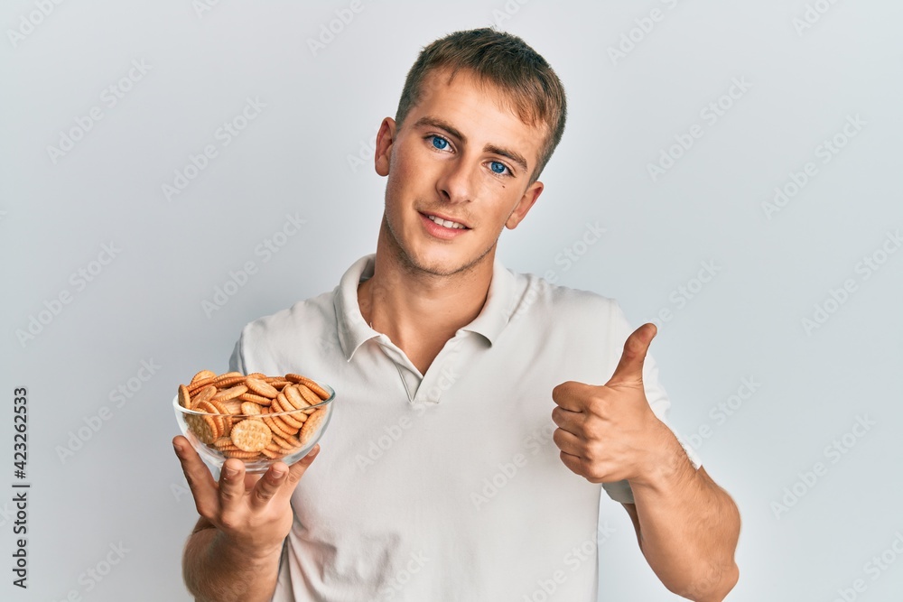 Young caucasian man holding salty biscuits bowl smiling happy and positive, thumb up doing excellent and approval sign Young caucasian man holding salty biscuits bowl smiling happy and positive, thumb up doing excellent and approval sign