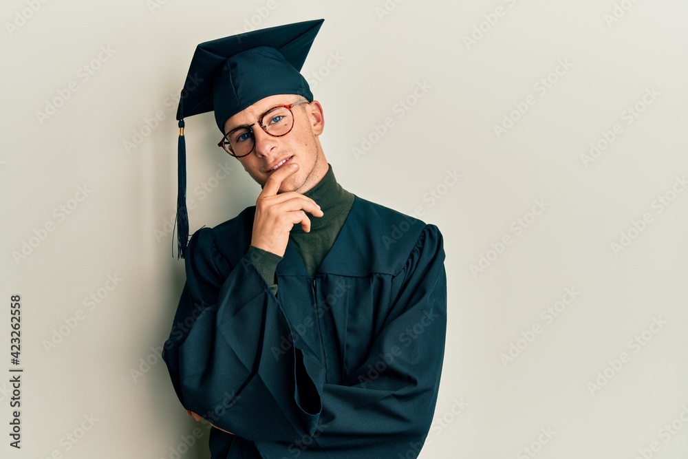 Young caucasian man wearing graduation cap and ceremony robe looking confident at the camera with smile with crossed arms and hand raised on chin. thinking positive. Young caucasian man wearing graduation cap and ceremony robe looking confident at the camera with smile with crossed arms and hand raised on chin. thinking positive.