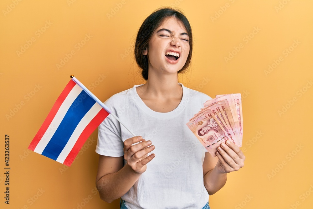 Young caucasian woman holding thailand flag and baht banknotes angry and mad screaming frustrated and furious, shouting with anger looking up. Young caucasian woman holding thailand flag and baht banknotes angry and mad screaming frustrated and furious, shouting with anger looking up.