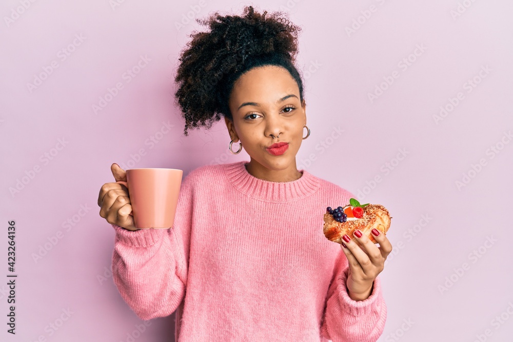 Young african american girl drinking a cup of coffee and eating sweet looking at the camera blowing a kiss being lovely and sexy. love expression. Young african american girl drinking a cup of coffee and eating sweet looking at the camera blowing a kiss being lovely and sexy. love expression.