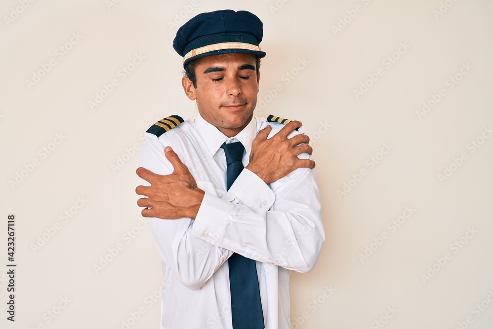 Young hispanic man wearing airplane pilot uniform hugging oneself happy and positive, smiling confident. self love and self care Young hispanic man wearing airplane pilot uniform hugging oneself happy and positive, smiling confident. self love and self care