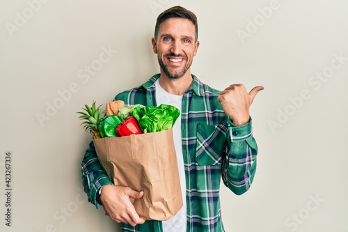Handsome man with beard holding paper bag with groceries pointing to the back behind with hand and thumbs up, smiling confident