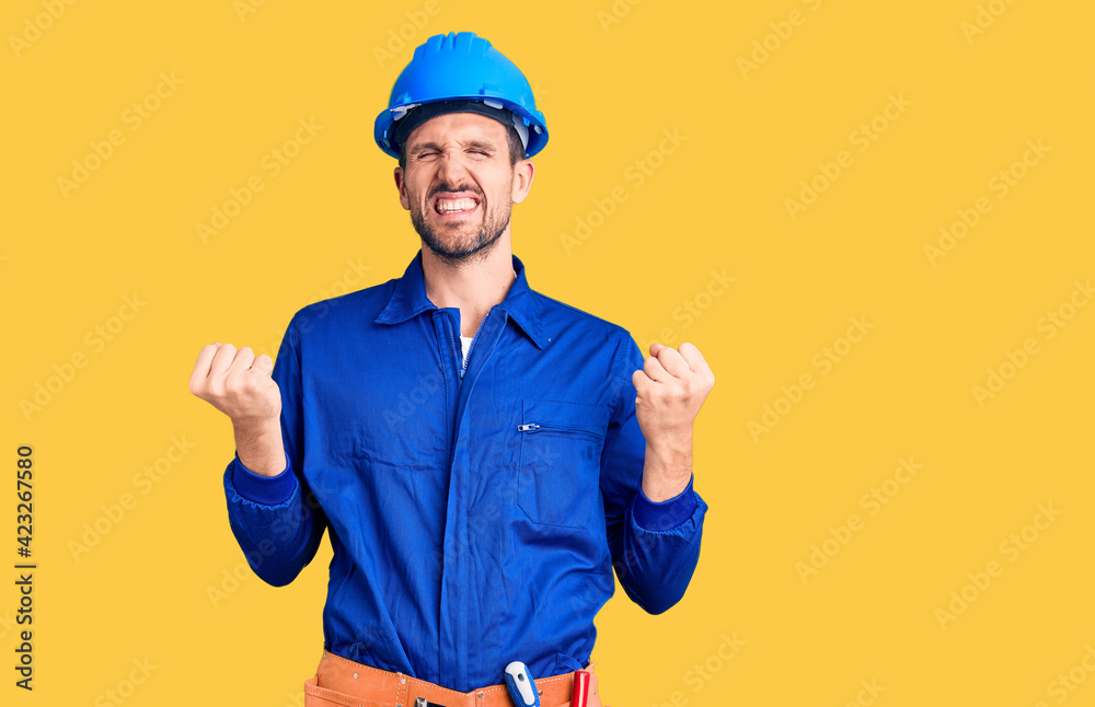 Young handsome man wearing worker uniform and hardhat very happy and excited doing winner gesture with arms raised, smiling and screaming for success. celebration concept. Young handsome man wearing worker uniform and hardhat very happy and excited doing winner gesture with arms raised, smiling and screaming for success. celebration concept.