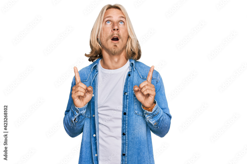 Obraz premium Caucasian man with blond long hair wearing casual denim jacket amazed and surprised looking up and pointing with fingers and raised arms.