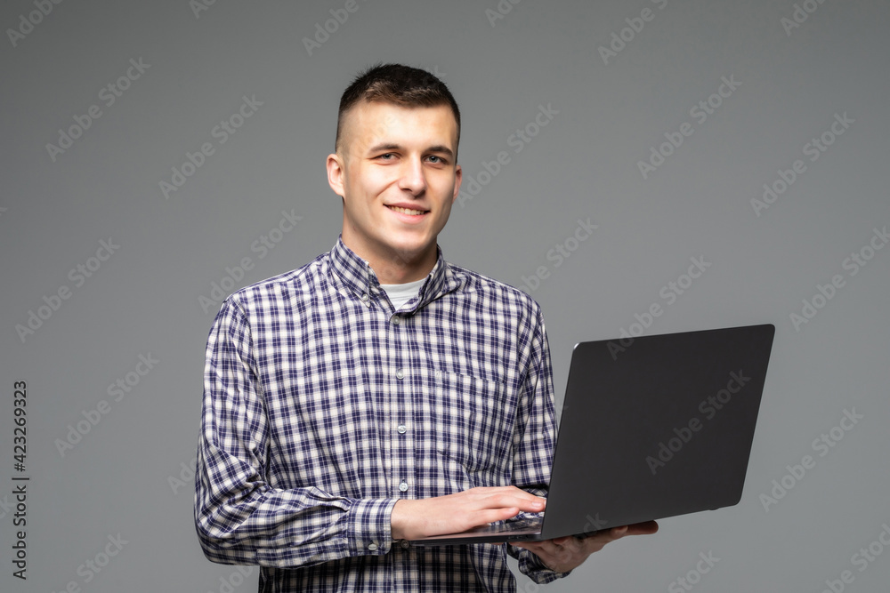 Checking his timetable. Confident young man holding laptop and typing while standing against grey background Checking his timetable. Confident young man holding laptop and typing while standing against grey background