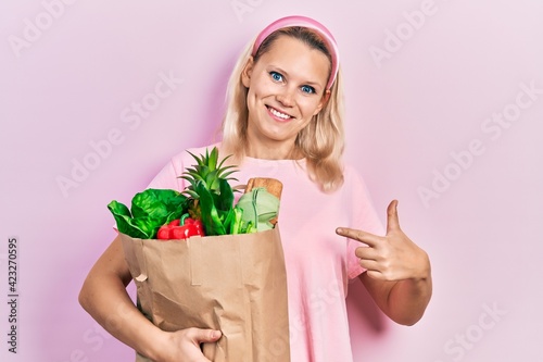 Beautiful caucasian blonde woman holding paper bag with groceries smiling happy pointing with hand and finger
