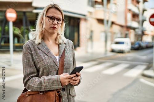 Young blonde businesswoman with serious expression using smartphone at the city.