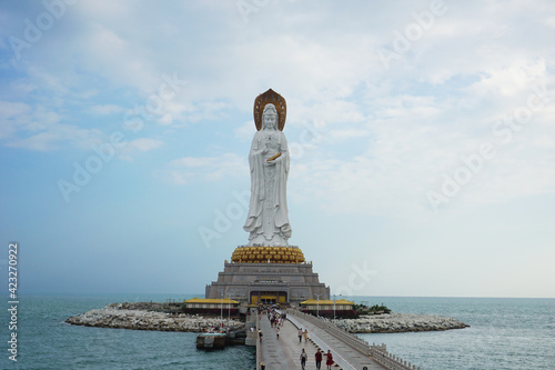  Statue of Guanyin at the Nanshan Buddhist Center on a cloudy day. A statue of the goddess Guanyin in the middle of the sea, pilgrims walking across the bridge. Sanya, Hainan, China