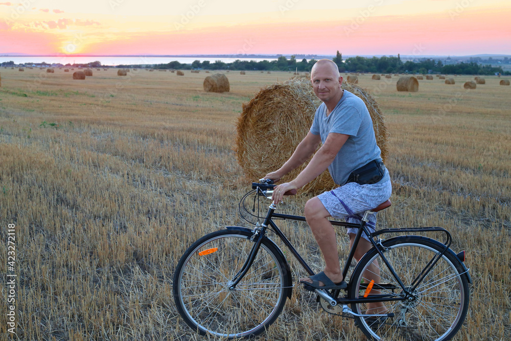 Obraz premium An adult man on a bicycle on a mowed field next to a straw walker at sunset. Sports and holiday activities in the countryside