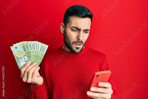 Young hispanic man using smartphone holding chilean pesos banknotes clueless and confused expression. doubt concept. photo