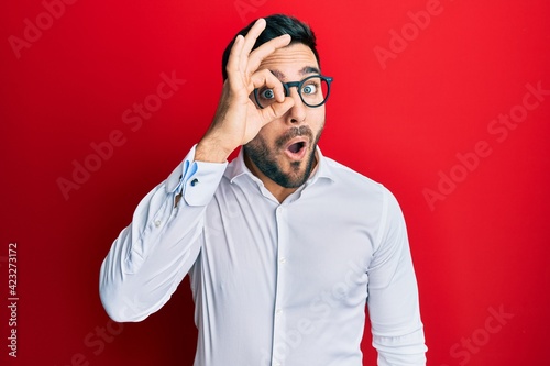Young hispanic businessman wearing shirt and glasses doing ok gesture shocked with surprised face, eye looking through fingers. unbelieving expression.