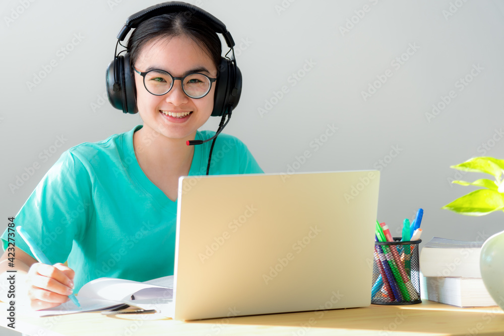Portrait young Asian female student with headphones glasses smile looking up a note on the book a teenage girl is happy to university internet distance study online class on a laptop computer at home Portrait young Asian female student with headphones glasses smile looking up a note on the book a teenage girl is happy to university internet distance study online class on a laptop computer at home