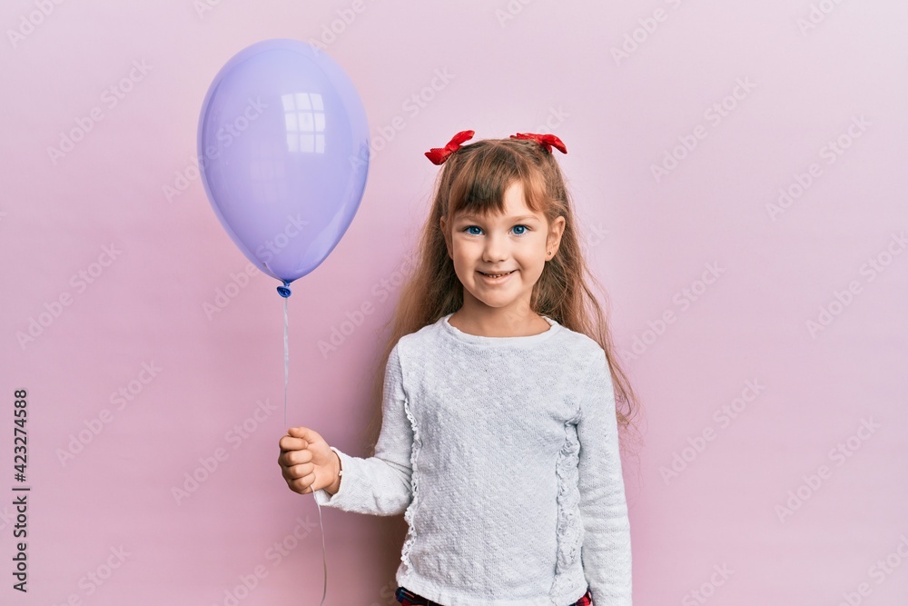 Little caucasian girl kid holding purple balloon looking positive and happy standing and smiling with a confident smile showing teeth