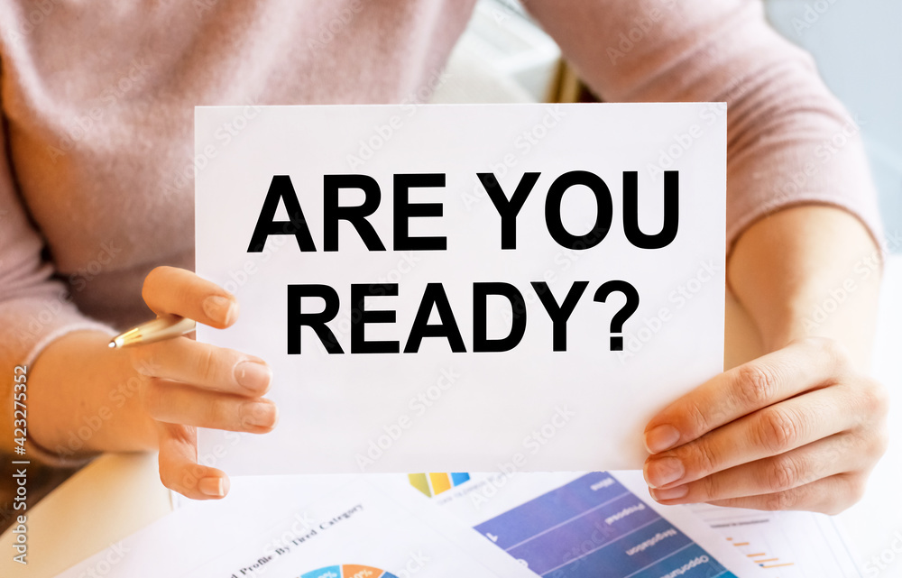 A girl in light clothes with a sign in her hands sits at the table. Lettering ARE YOU READY. Content lettering corresponds to business concept. A girl in light clothes with a sign in her hands sits at the table. Lettering ARE YOU READY. Content lettering corresponds to business concept.