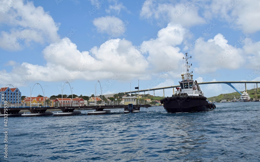 Pilot tug boat passing Queen Emma Bridge pontoon bridge across St. Anna Bay in Willemstad Curaçao; partially open with the colorful buildings of Otrobanda and the Queen Juliana Bridge in the backgroun