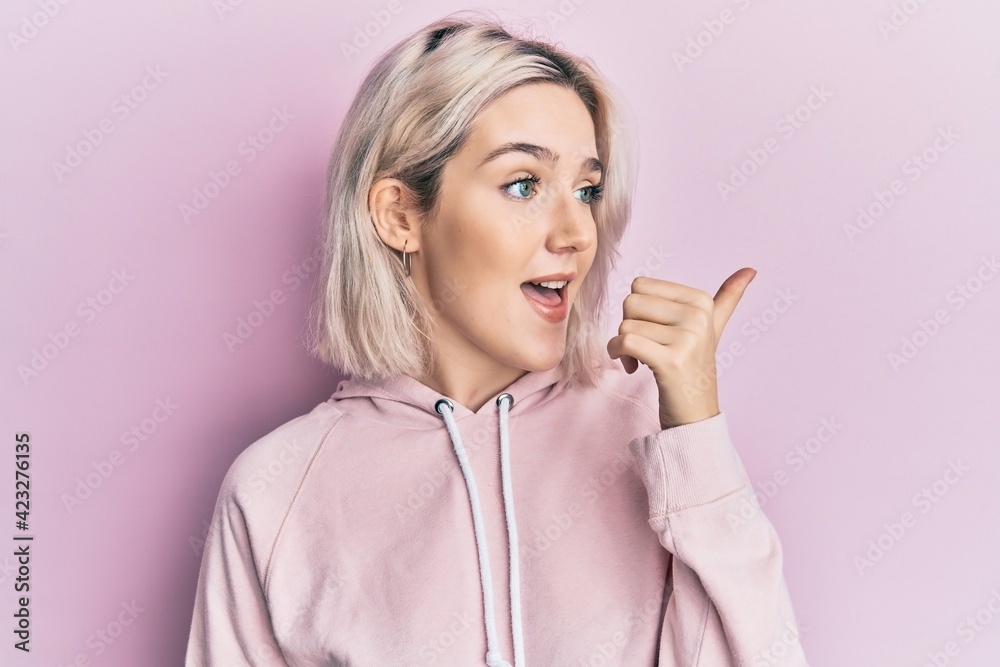 Young blonde girl wearing casual clothes pointing thumb up to the side smiling happy with open mouth Young blonde girl wearing casual clothes pointing thumb up to the side smiling happy with open mouth