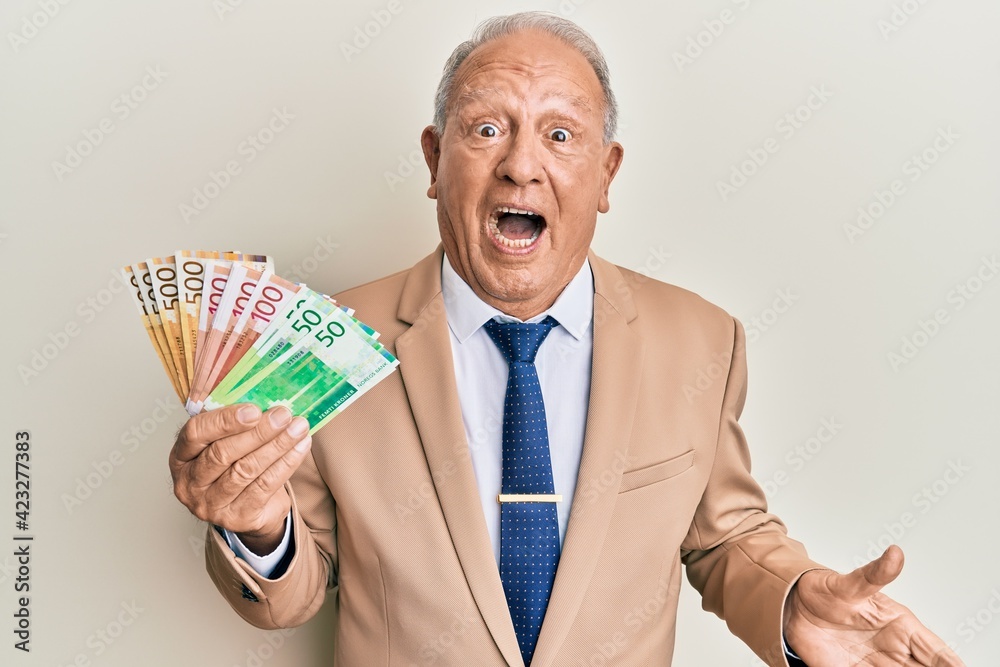 Senior caucasian man holding norwegian krone banknotes celebrating achievement with happy smile and winner expression with raised hand Senior caucasian man holding norwegian krone banknotes celebrating achievement with happy smile and winner expression with raised hand