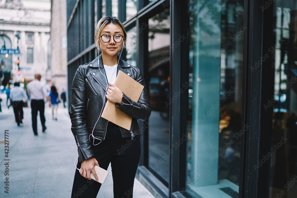 Half length portrait of millennial blogger in eyeglasses holding education sketchbook looking at camera while listening music podcast, Chinese female in electronic headphones using cellphone app