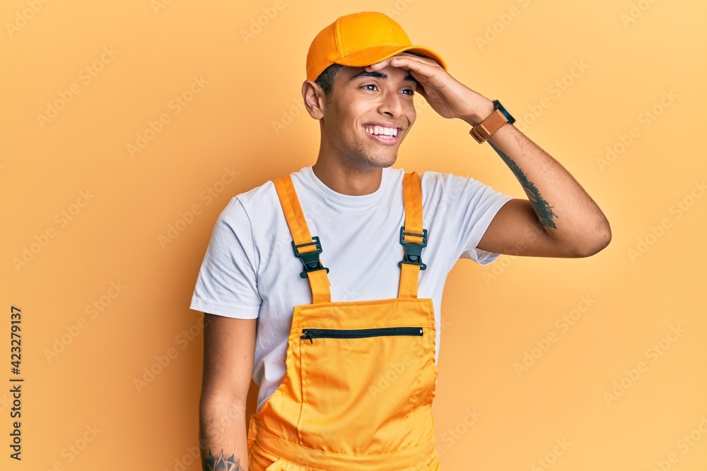 Young handsome african american man wearing handyman uniform over yellow background very happy and smiling looking far away with hand over head. searching concept. Young handsome african american man wearing handyman uniform over yellow background very happy and smiling looking far away with hand over head. searching concept.