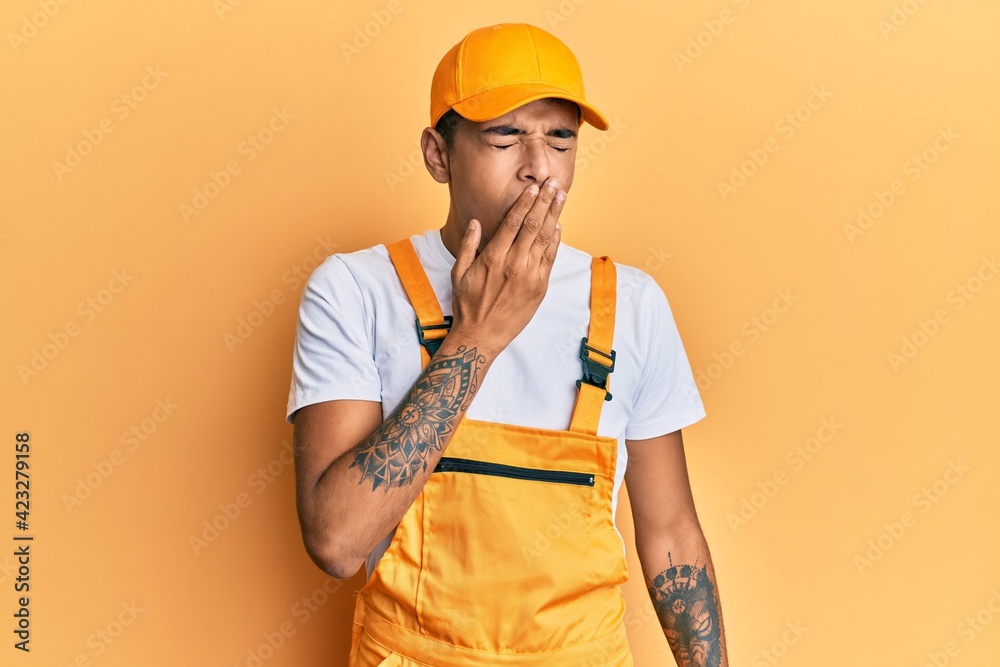 Young handsome african american man wearing handyman uniform over yellow background bored yawning tired covering mouth with hand. restless and sleepiness.
