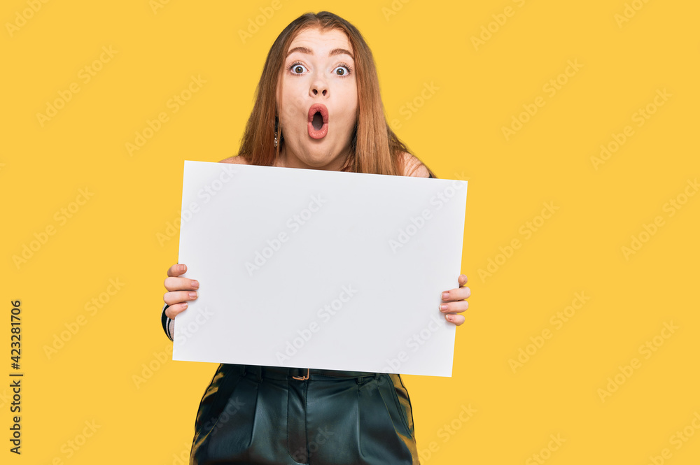 Young beautiful redhead woman holding blank empty banner scared and amazed with open mouth for surprise, disbelief face Young beautiful redhead woman holding blank empty banner scared and amazed with open mouth for surprise, disbelief face