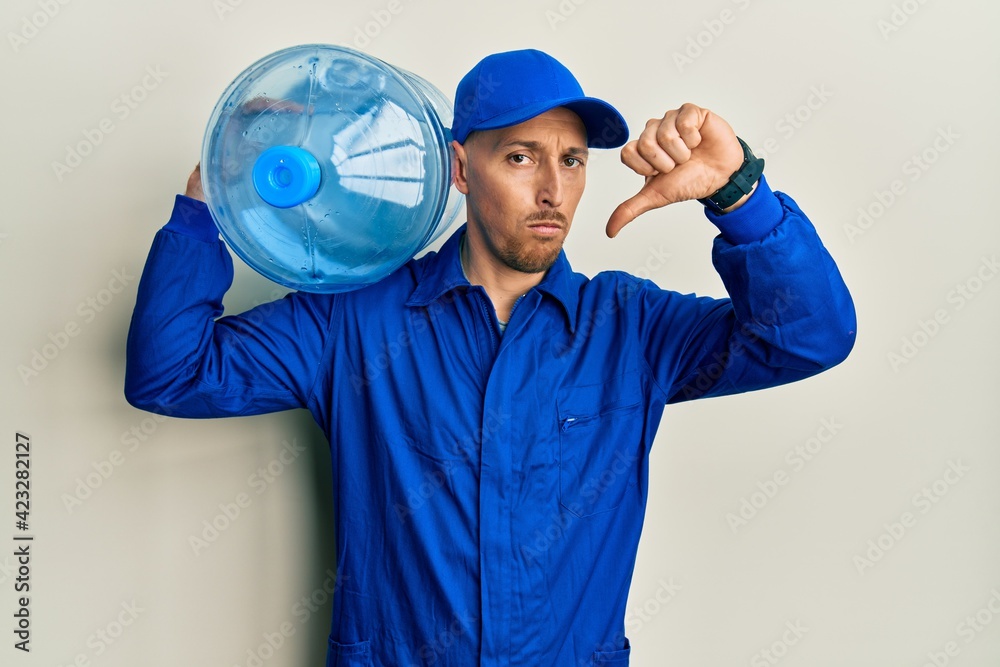 Bald courier man with beard holding a gallon bottle of water for delivery with angry face, negative sign showing dislike with thumbs down, rejection concept