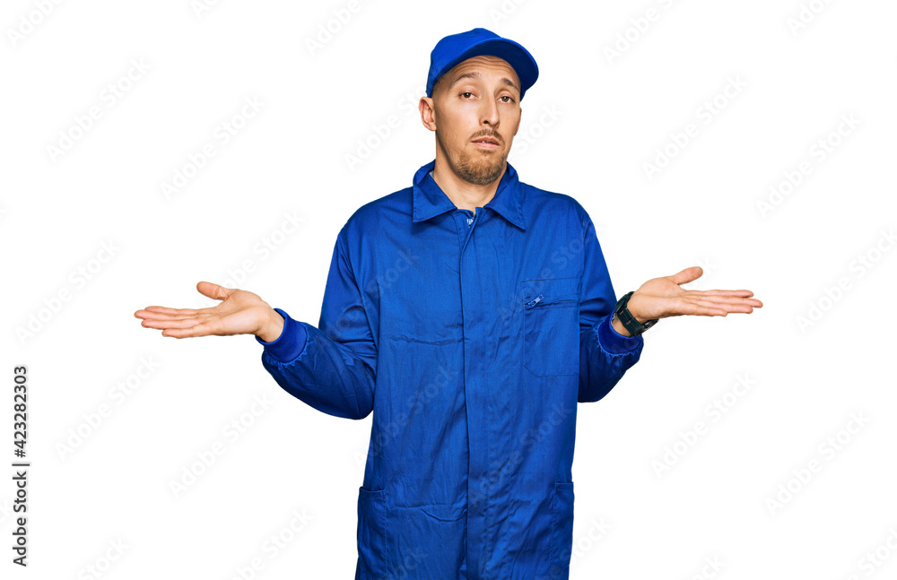 Bald man with beard wearing builder jumpsuit uniform clueless and confused expression with arms and hands raised. doubt concept.