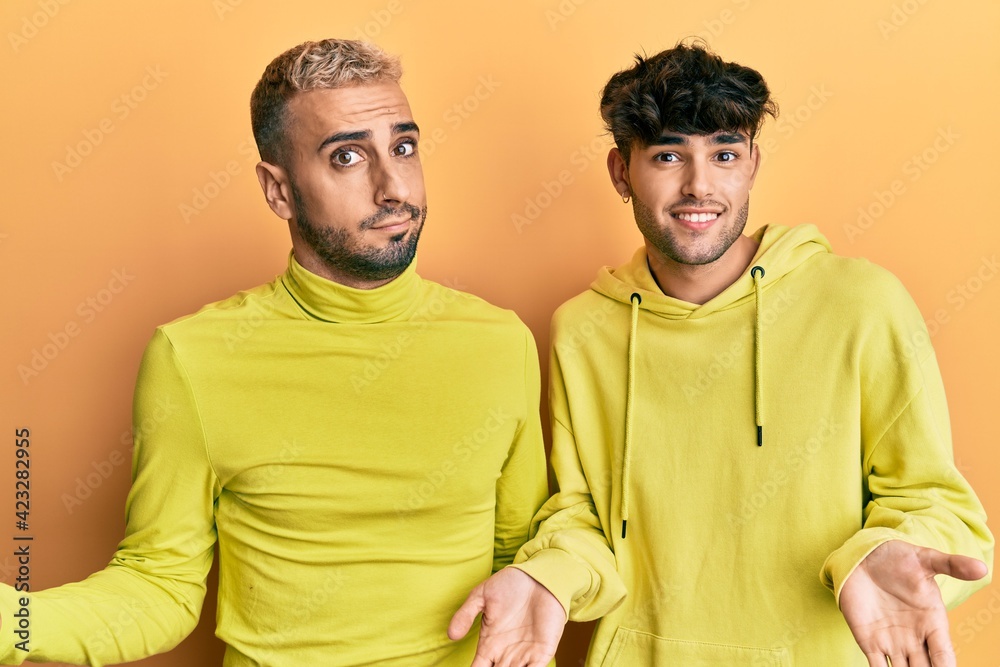 Homosexual gay couple standing together wearing yellow clothes clueless and confused expression with arms and hands raised. doubt concept. Homosexual gay couple standing together wearing yellow clothes clueless and confused expression with arms and hands raised. doubt concept.