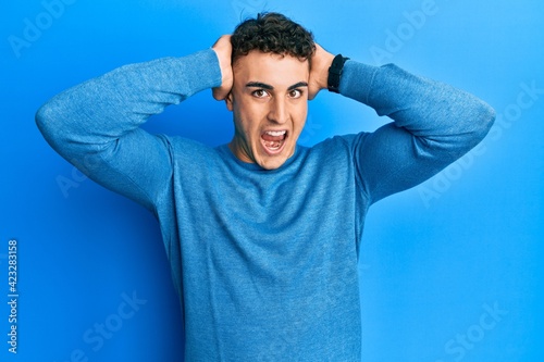 Hispanic young man wearing casual winter sweater crazy and scared with hands on head, afraid and surprised of shock with open mouth