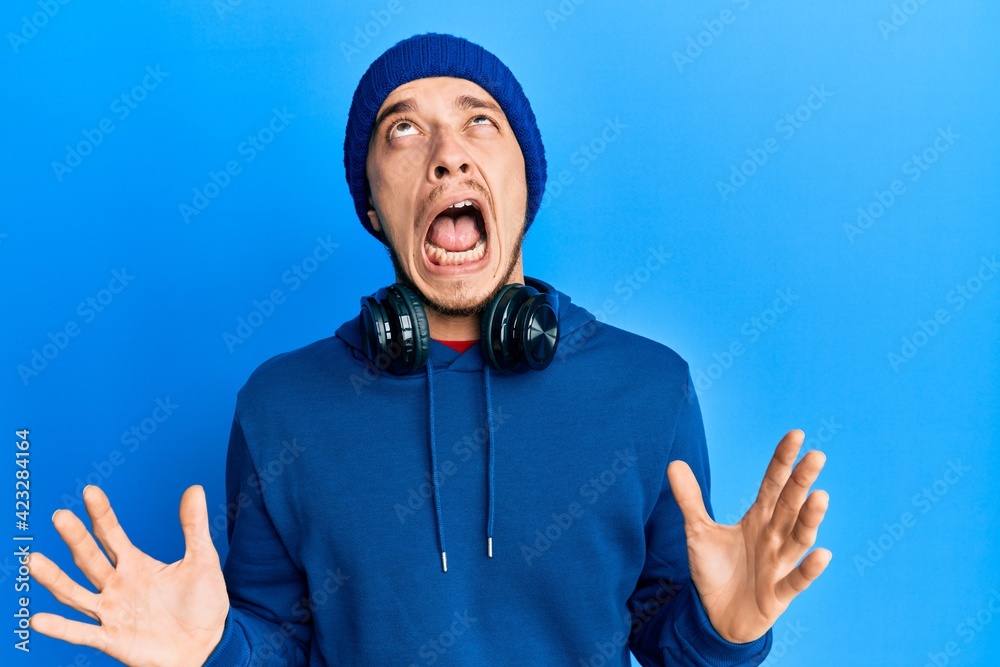 Naklejka premium Hispanic young man wearing sweatshirt and headphones crazy and mad shouting and yelling with aggressive expression and arms raised. frustration concept.