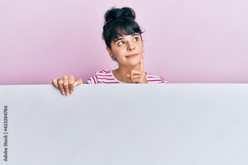 Young hispanic girl holding blank empty banner serious face thinking about question with hand on chin, thoughtful about confusing idea Young hispanic girl holding blank empty banner serious face thinking about question with hand on chin, thoughtful about confusing idea
