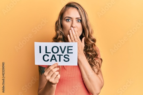 Young blonde girl holding paper with i love cats phrase covering mouth with hand, shocked and afraid for mistake. surprised expression